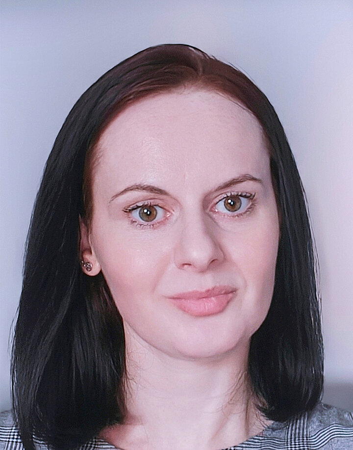 Uśmiechnięta młoda kobieta o słowiańskich rysach i ciemnych długich włosach. Nosi sukienkę w kratę.