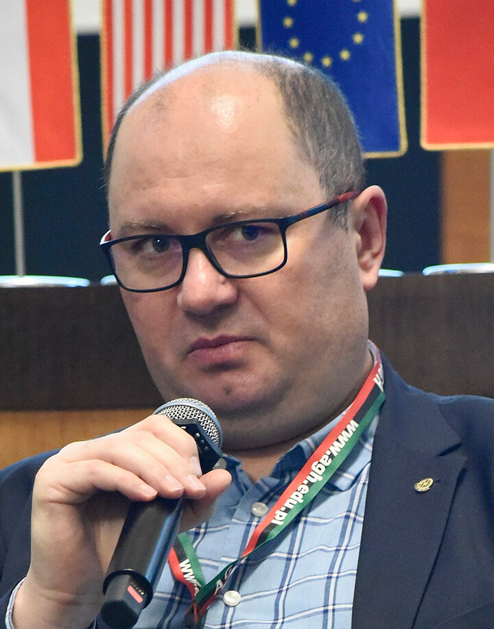 Łysiejący mężczyzna w średnim wieku, w okularach z ciemnymi oprawkami. Nosi koszyulę w paski oraz grantową marynarkę.