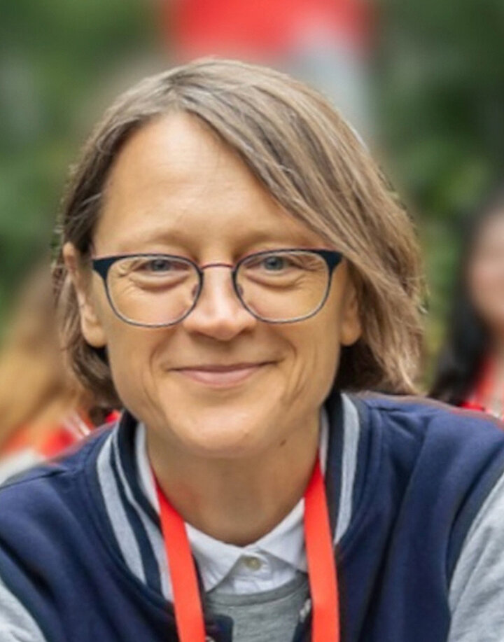 Uśmiechnięta kobieta w średnim wieku o półdługich blond włosach. Nosi okulary oraz sportową kurtrkę.