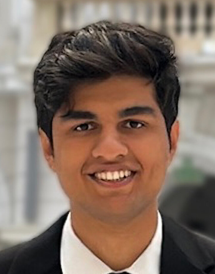 Uśmiechnięty młody mężczyzna o hiduskich rysach i bujnych czarnych włosach. Nosi białą koszulę i czarną marynarkę.