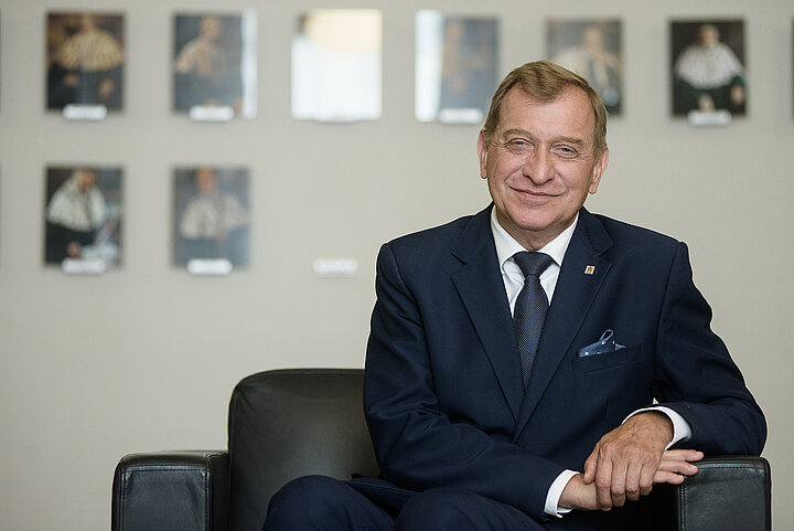 Starszy mężczyzna o blond włosach w granatowym garniturze siedzi w prostym fotelu i uśmiecha się do obserwatora.