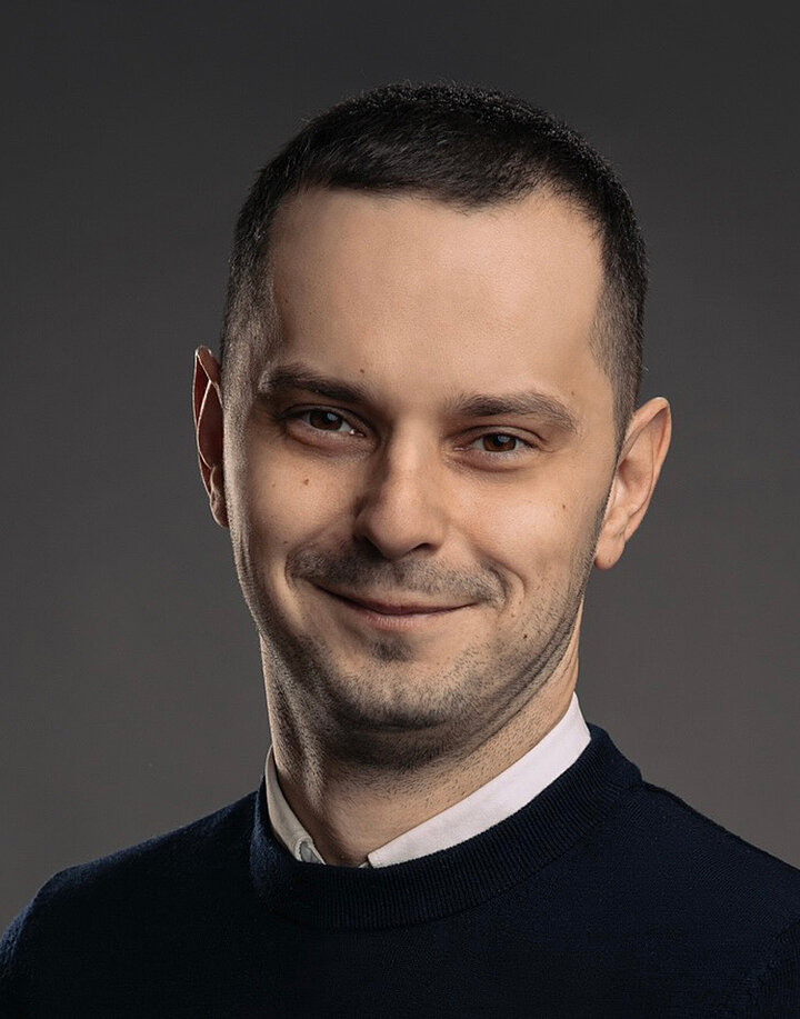 Młody, usmiechnięty mężczyzna w białej koszuli i czarnym swetrze.