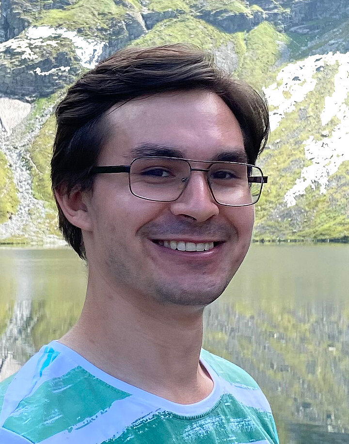 Młody, umiechnięty mężczyzna o słowiańskich rysach. Nosi okulary z cienkimi oprawkami. Ma na sobie biało-zielony t-shirt. W tle panorama Tatr.