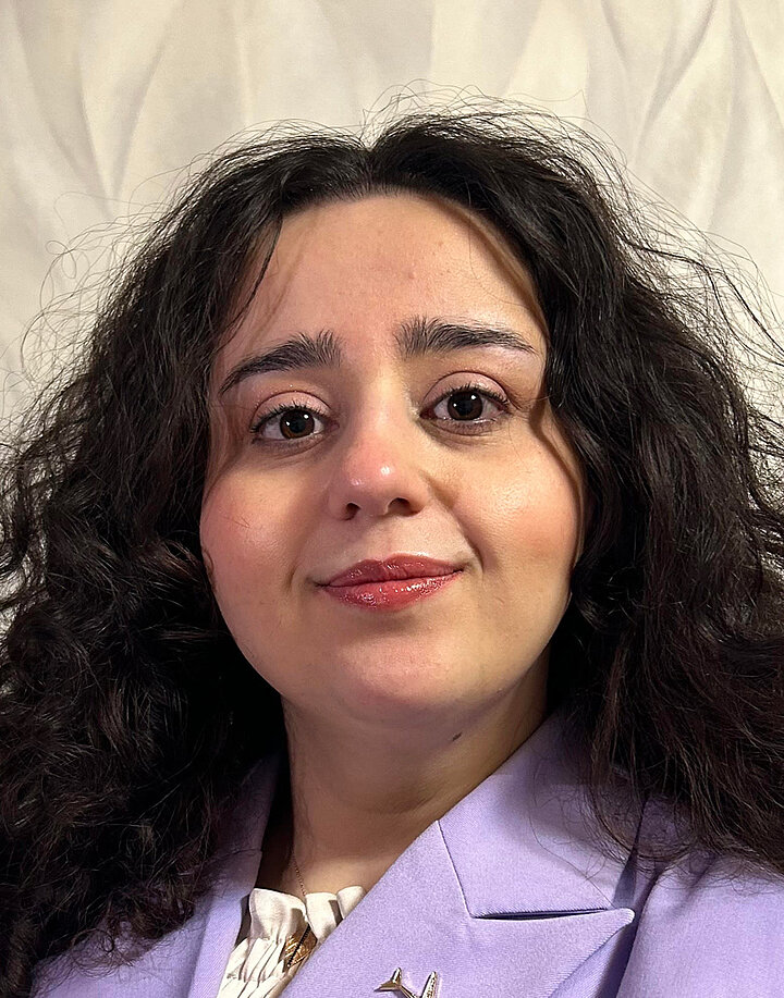 Młoda uśmiechnięta kobieta o bliskowshodnich rysach twarzy i ciemnych długich włosach. Nosi fioletowy żakiet.