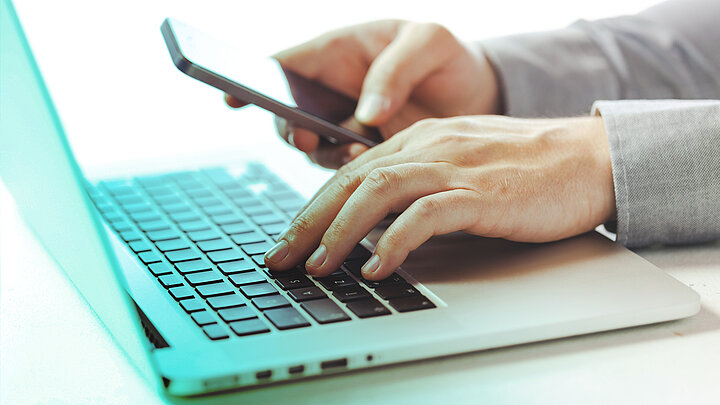 Zbliżenie na dłonie mężczyzny nad klawiaturą. W prawej trzyma smartfon