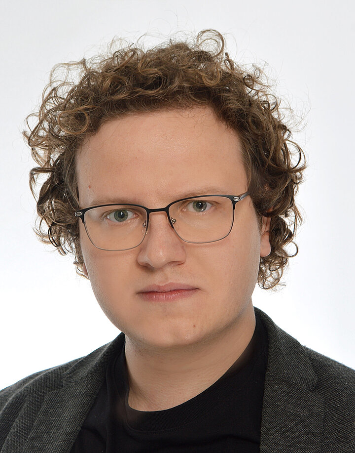 Młody mężczyzna o europelskich rysach i rozwianymi blnd włosami. Nosi okulary i ciemna marynarkę.