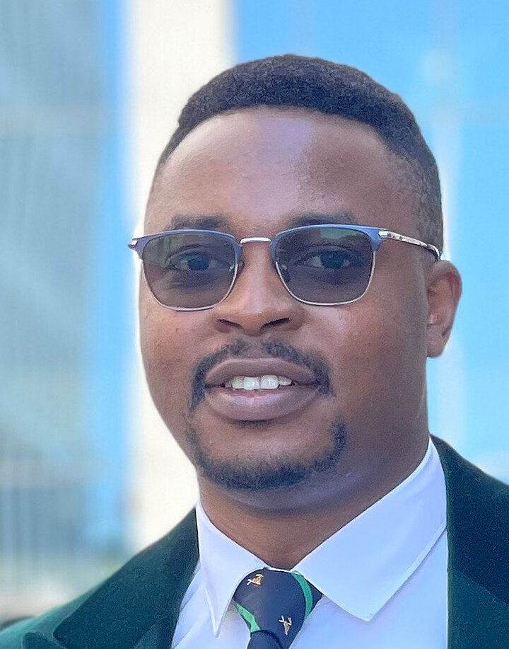 Uśmiechnięty mężczyzna o afrykańskich rysach w ciemnych okularach korekcyjnych, noszcy wąsy i brodę. Ma ciemny garnitur.