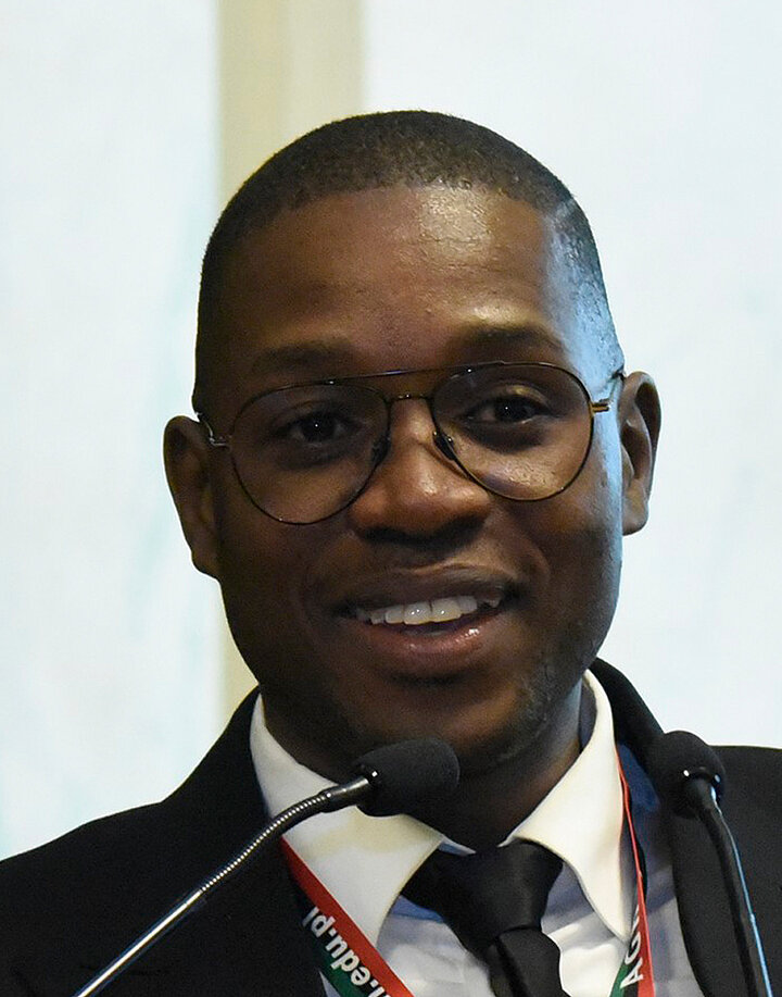 Krótko ostrzyżony, uśmiechnięty mężczyzna o afrykańskich rysach. Nosi okulary w ceimnych oprawkach oraz czarną marynarkę.